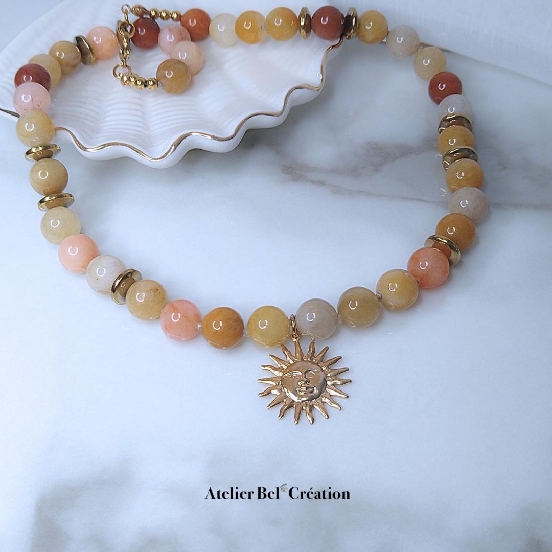 Peut inclure: Un collier de perles avec un pendentif soleil. Le collier est composé de perles pêche, jaunes et marron avec des accents dorés. Le pendentif soleil est doré avec un visage souriant. Le texte "Atelier Bel Création" est en bas.